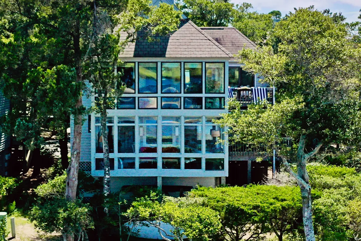 A striking house with large glass windows sits surrounded by lush greenery. The inviting balcony overlooks nature, with ample outdoor seating for relaxation and gatherings, conveying a peaceful, secluded vibe.