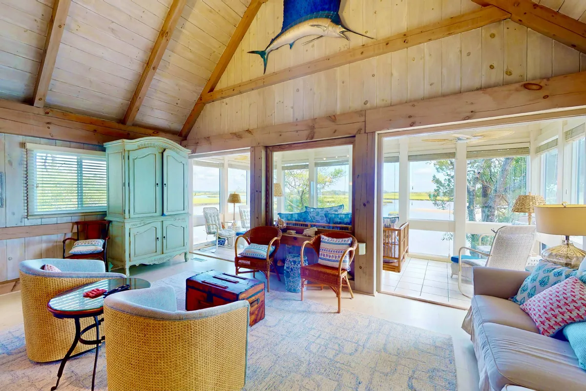 A warm, casual living area with expansive windows framing the outdoors, a mounted fish accent on the ceiling, and a comfortable seating arrangement ideal for gatherings. The room’s layout and decor hint at summer afternoons spent by the windows and cozy evenings with friends, with sunset light streaming in.