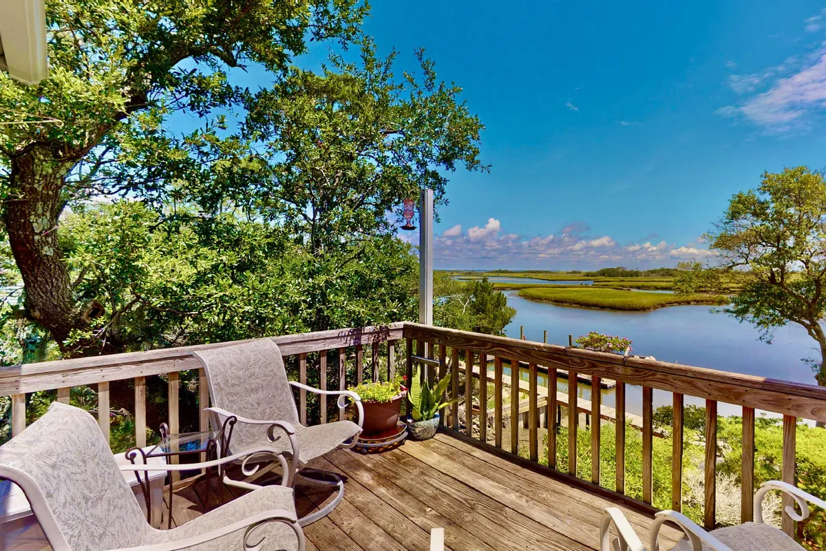 The image showcases a sun-dappled wooden deck overlooking serene Bald Head Creek, surrounded by lush greenery. Comfortable outdoor seating invites guests to relax, with tranquil water views and live oaks framing the scene—perfect for watching wildlife or savoring quiet moments in nature.