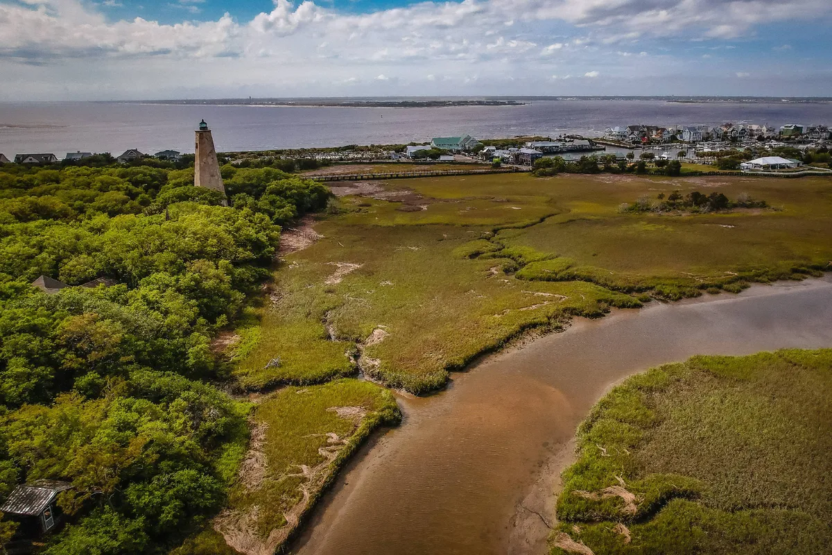 A picturesque vista featuring a lighthouse set against marshland and a shimmering waterway, with a town visible in the distance. The expansive marsh terrain encourages exploration, birdwatching, and family hikes.