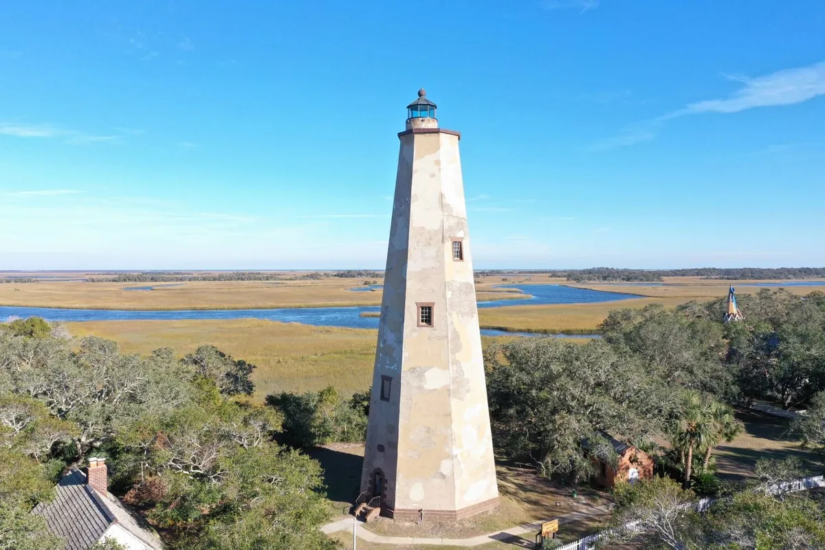 This image features the iconic Old Baldy Lighthouse rising above surrounding marshland, with the river and lush vegetation providing a tranquil natural backdrop. The composition emphasizes the lighthouse as a historic landmark and the expansive, serene setting of Bald Head Island.