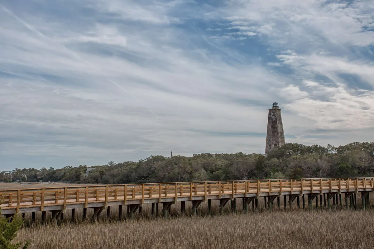A tranquil boardwalk winds its way through marshland toward a distant lighthouse. The path is bordered by wild grasses and offers scenic views, inviting exploration and wildlife spotting.
