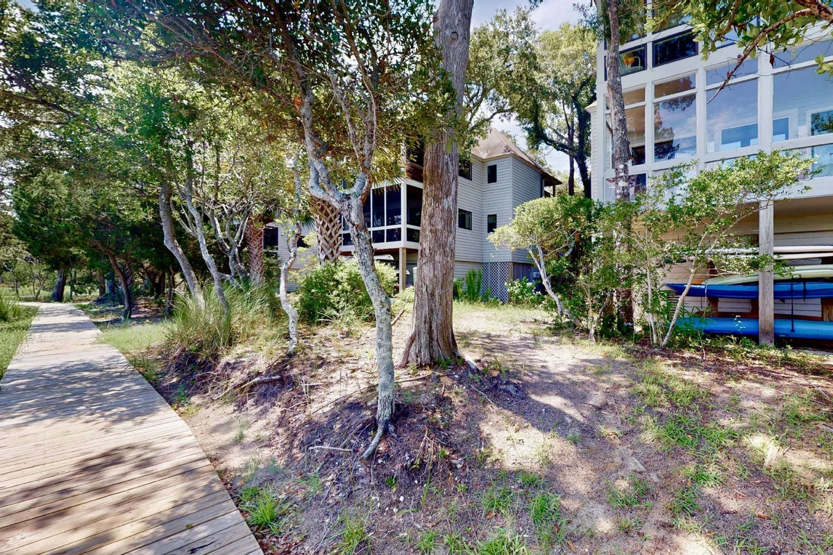 A relaxed exterior scene featuring a wooden pathway bordered by trees, houses with large windows, and visible kayaks, capturing the boardwalk leading out to the water and the community’s natural setting.