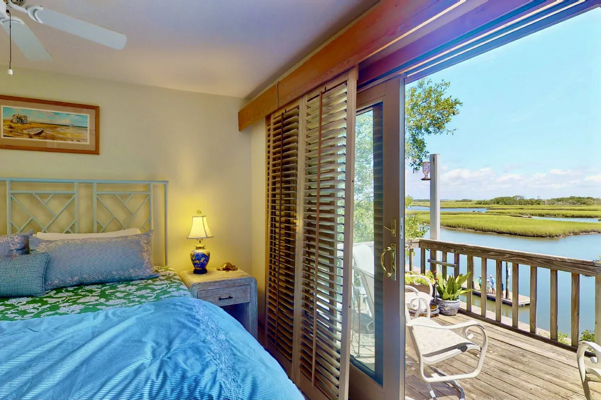 A serene bedroom with a bed featuring blue and green bedding, a large window framing views of the marsh, and a nightstand with a lamp and decorative accents. The space feels airy and peaceful, inviting relaxation and a connection to nature.