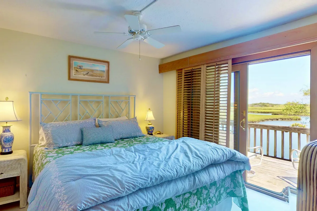 A calm and inviting bedroom with floral bedding, a large sliding glass door opening to a scenic view, decorative lamps, and framed artwork. Natural light flows into the room, enhancing its restful ambiance.