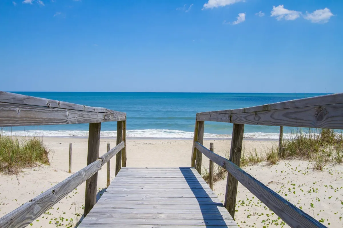 A wooden boardwalk weaves through dune grass, leading to the sandy beach with gentle ocean waves in the background. The expansive sky and natural landscape create an inviting path for walks and exploration.