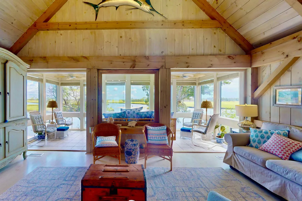 A spacious living area with large windows offering sweeping views of the marsh and Bald Head Creek. The room features wooden beams and walls, a comfortable furniture arrangement for group gatherings, and a whimsical decorative fish mounted on the wall. Ample natural light floods the space, making it inviting for relaxing with a book, socializing, or sunset watching by the fireplace.