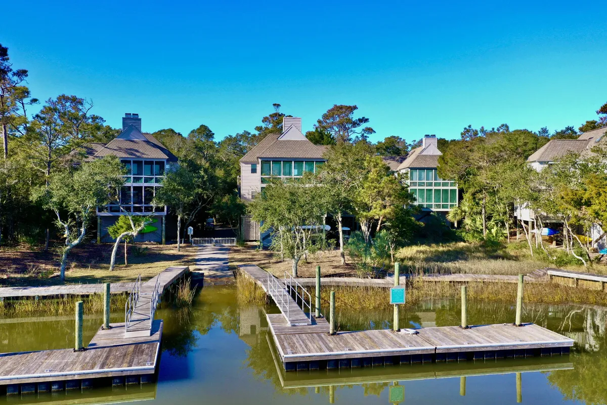 A peaceful exterior view of a dock leading out over the water, bordered by houses and shaded by trees. The scene suggests easy access to nature and opportunities for boating, fishing, or relaxing while wildlife watching.