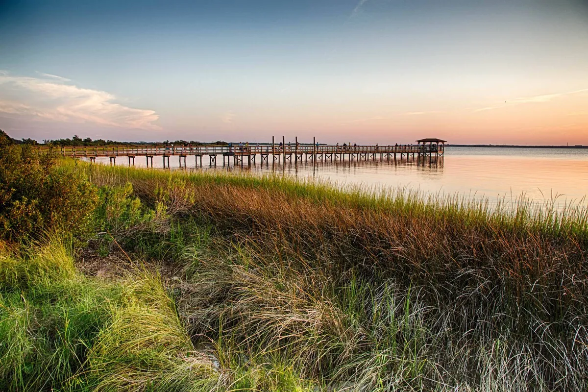 A wooden pier stretches into calm water beneath a colorful sunset sky, bordered by a grassy shoreline. The scene is peaceful and inviting, evoking moments of togetherness by the water.