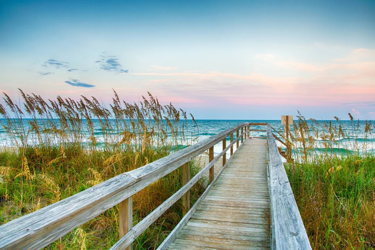 A wooden boardwalk stretches toward ocean waves, bordered by dune grass and set against a vibrant sky. The scene evokes feelings of tranquility, inviting visitors to stroll along the shore, take in sunset views, and experience the island’s unspoiled coastal landscapes.